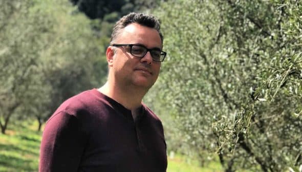 Man wearing glasses standing in an olive grove with trees in the background. - Olive Oil Times