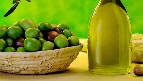 A basket filled with green and brown olives next to a bottle of olive oil on a table. - Olive Oil Times