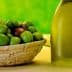 A basket filled with green and brown olives next to a bottle of olive oil on a table. - Olive Oil Times