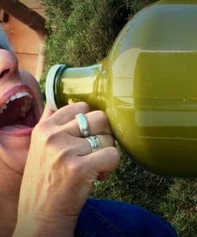 Woman with gray hair drinking from a large green glass bottle outdoors. - Olive Oil Times