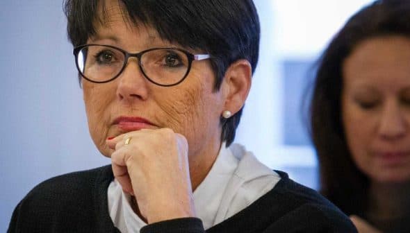 Woman with short black hair and glasses sitting at a table during a meeting, looking thoughtful. - Olive Oil Times