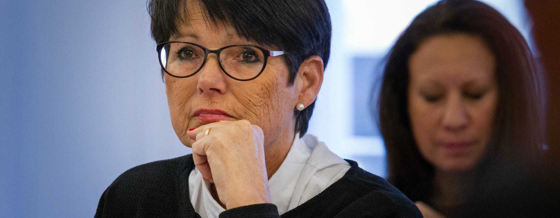 Woman with short black hair and glasses sitting at a table during a meeting, looking thoughtful. - Olive Oil Times