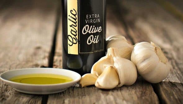 Bottle of garlic extra virgin olive oil next to garlic bulbs and a small dish of olive oil. - Olive Oil Times