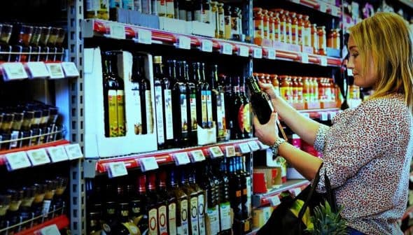 A woman examining a bottle of olive oil while shopping in a grocery store aisle filled with various oils. - Olive Oil Times