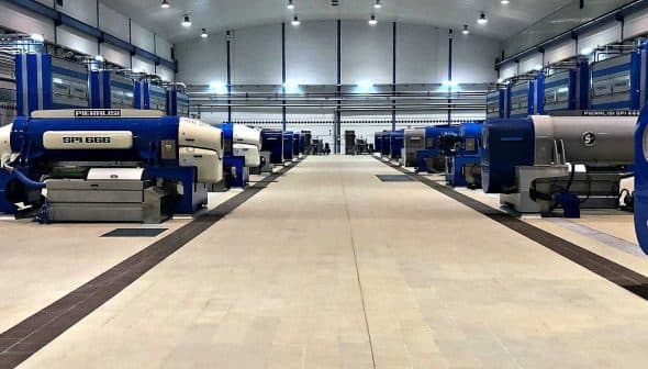 Row of industrial olive oil processing machines in a large facility with blue and gray colors. - Olive Oil Times