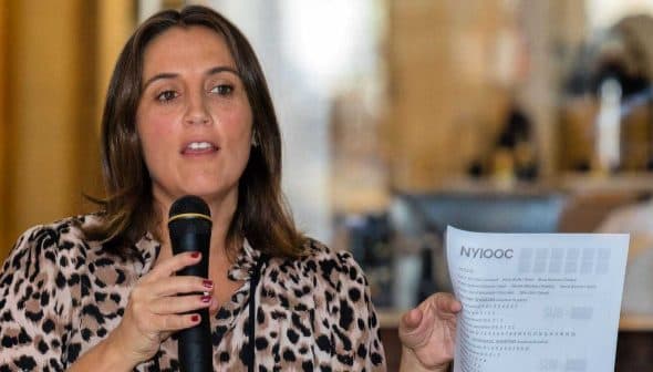 Woman holding a microphone and a sheet of paper while speaking at an event. - Olive Oil Times