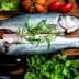 Two raw fish placed on a wooden cutting board surrounded by fresh vegetables and herbs. - Olive Oil Times