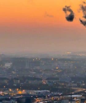 Smoke rising from a tall industrial chimney against a sunset sky with city skyline in the background. - Olive Oil Times