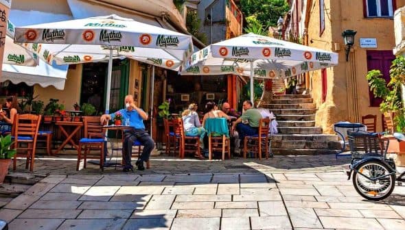 Outdoor café scene featuring patrons seated under large umbrellas in a narrow street. - Olive Oil Times