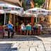 Outdoor café scene featuring patrons seated under large umbrellas in a narrow street. - Olive Oil Times