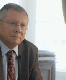 A man in a suit sitting at a desk with a serious expression, wearing glasses and a tie. - Olive Oil Times
