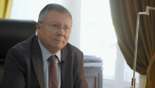 A man in a suit sitting at a desk with a serious expression, wearing glasses and a tie. - Olive Oil Times