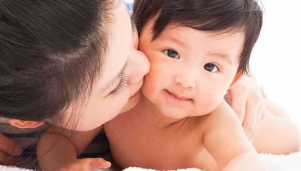A mother kissing her baby on a soft surface, both looking at the camera. - Olive Oil Times