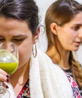 Woman holding a glass of green liquid while tasting olive oil at an event with two other women in the background. - Olive Oil Times