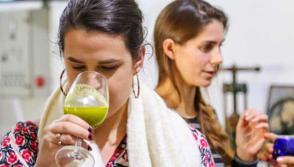 Woman holding a glass of green liquid while tasting olive oil at an event with two other women in the background. - Olive Oil Times