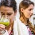 Woman holding a glass of green liquid while tasting olive oil at an event with two other women in the background. - Olive Oil Times