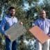 Two men standing outdoors holding samples of textured materials in front of an olive tree. - Olive Oil Times