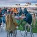 Group of people sitting and standing at a rooftop bar with tables and chairs. - Olive Oil Times