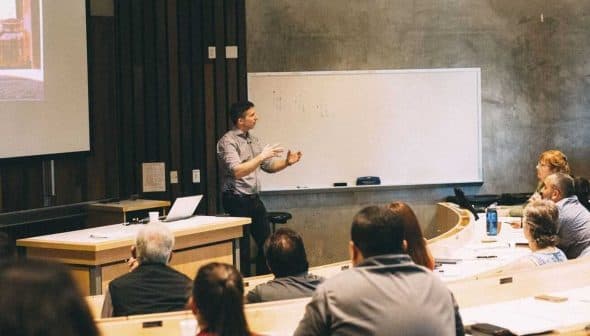 A speaker presenting in front of an audience in a classroom with a whiteboard. - Olive Oil Times