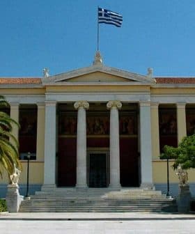 Facade of the University of Athens building with Greek flag and palm trees in front. - Olive Oil Times