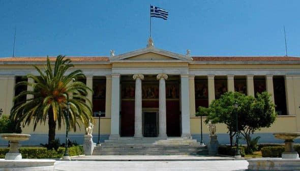 Facade of the University of Athens building with Greek flag and palm trees in front. - Olive Oil Times