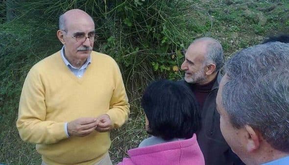 A man wearing a yellow sweater speaking to a small group of people outdoors. - Olive Oil Times