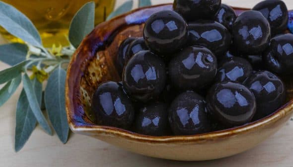 A ceramic bowl filled with glossy black olives, accompanied by olive oil and olive leaves. - Olive Oil Times
