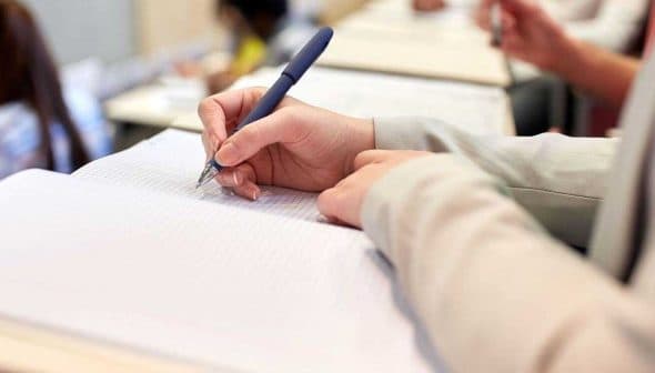 A person holding a pen and writing in a notebook during a classroom setting. - Olive Oil Times