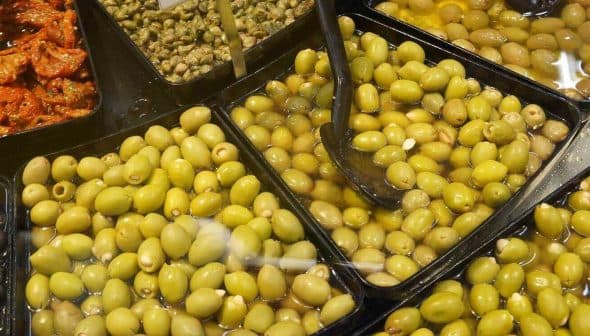 Multiple trays containing different types of olives, including green and black varieties, with serving utensils. - Olive Oil Times