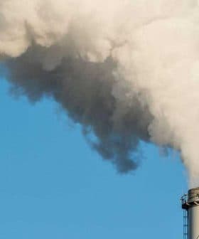 Two industrial smoke stacks releasing white vapor into a clear blue sky. - Olive Oil Times