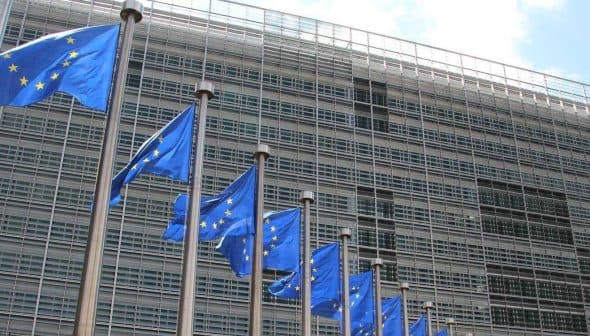 Multiple European Union flags displayed in front of a modern glass building. - Olive Oil Times