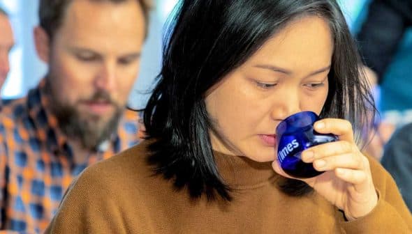 Woman with black hair holding a blue glass to her lips while tasting. - Olive Oil Times