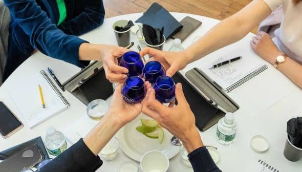 Four individuals toasting with blue glasses at a round table during a meeting. - Olive Oil Times