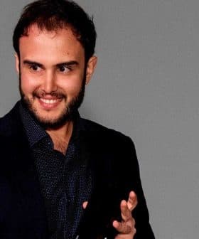 Two men standing on stage, one smiling and clapping while the other speaks. - Olive Oil Times