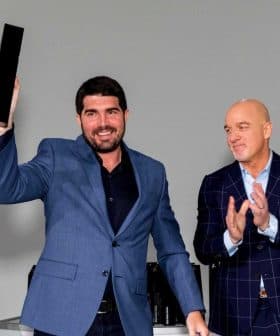 A man in a blue suit holding an award while two others applaud in the background. - Olive Oil Times