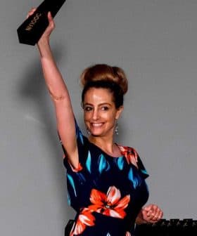 Woman in a floral dress holding an award above her head during an event with two people clapping in the background. - Olive Oil Times