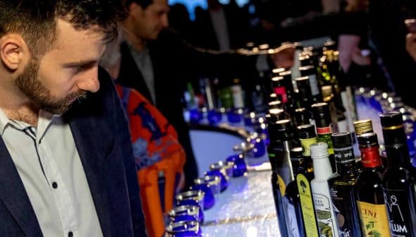 Man in a dark suit looking at a row of various bottles on a bar at an event. - Olive Oil Times