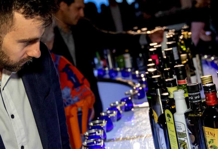 Man in a dark suit looking at a row of various bottles on a bar at an event. - Olive Oil Times