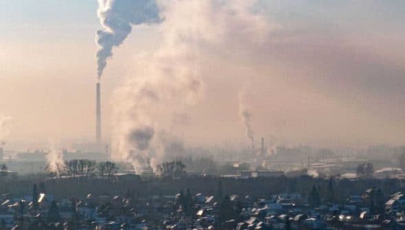 Smoke rising from industrial chimneys in an urban area with a hazy sky. - Olive Oil Times