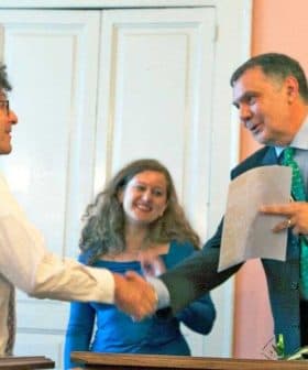 Two individuals shaking hands during an award ceremony with attendees in the background. - Olive Oil Times