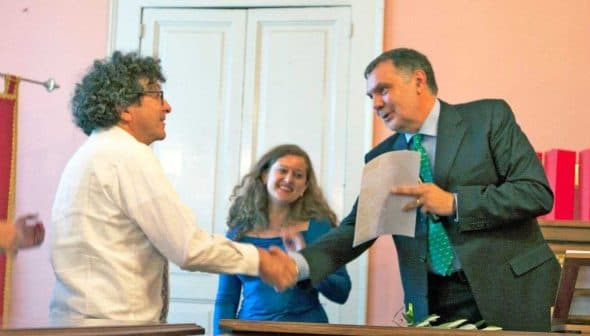Two individuals shaking hands during an award ceremony with attendees in the background. - Olive Oil Times