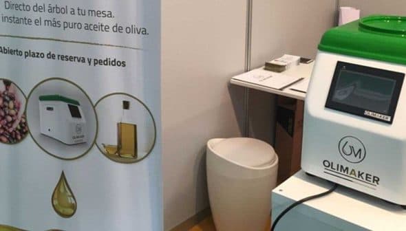 Olimaker machine for producing olive oil displayed at an event with informational banner. - Olive Oil Times
