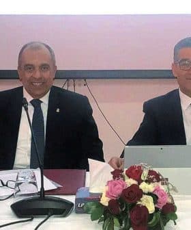 Four individuals seated at a conference table with a flag and floral arrangement in front of them. - Olive Oil Times