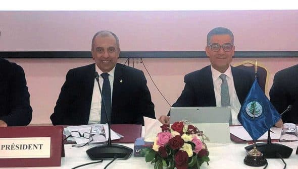Four individuals seated at a conference table with a flag and floral arrangement in front of them. - Olive Oil Times