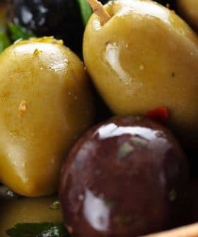A close-up of mixed green and black olives garnished with herbs in a wooden bowl. - Olive Oil Times