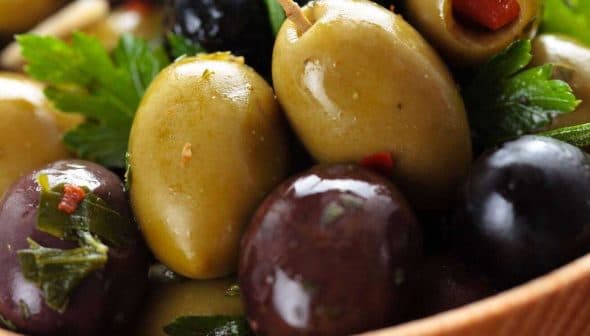 A close-up of mixed green and black olives garnished with herbs in a wooden bowl. - Olive Oil Times