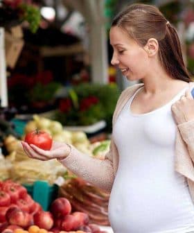 Pregnant woman examining a tomato while shopping at a farmers market with various fruits and vegetables around her. - Olive Oil Times