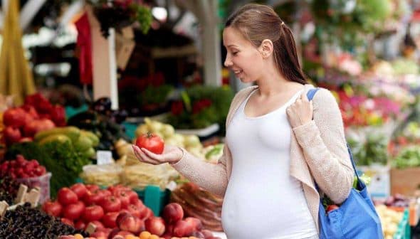Pregnant woman examining a tomato while shopping at a farmers market with various fruits and vegetables around her. - Olive Oil Times