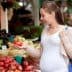 Pregnant woman examining a tomato while shopping at a farmers market with various fruits and vegetables around her. - Olive Oil Times