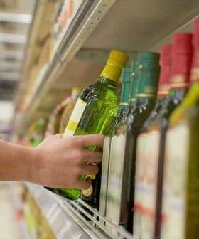 Individual reaching for a green bottle of olive oil on a supermarket shelf among other oil bottles. - Olive Oil Times